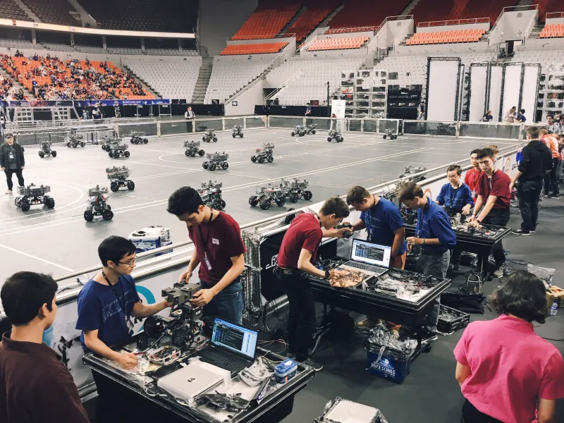 Teen teams tuning wheeled robots at pit tables beside a large arena competition floor