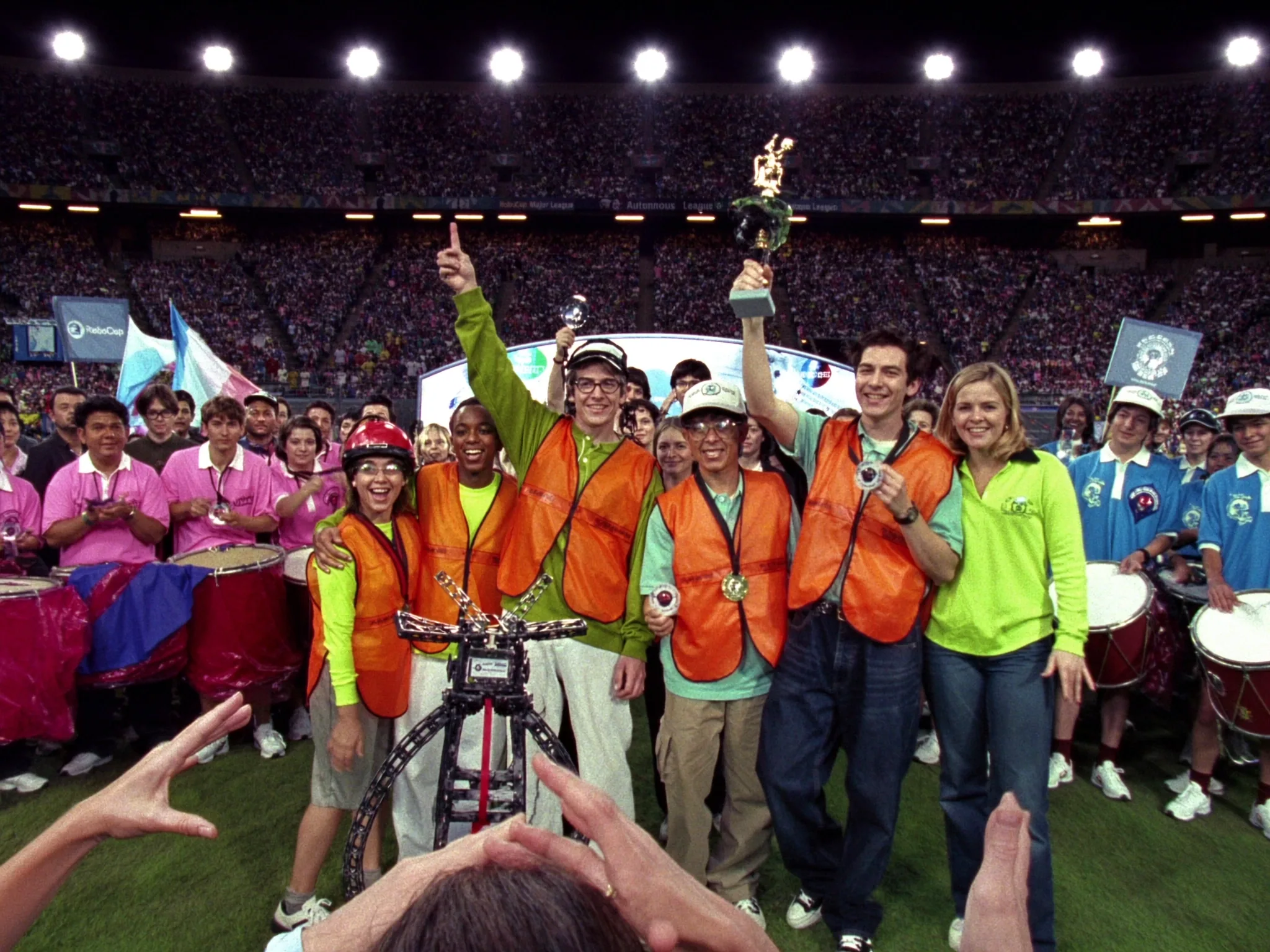 Team in orange vests raising a trophy beside their robot on a stadium field