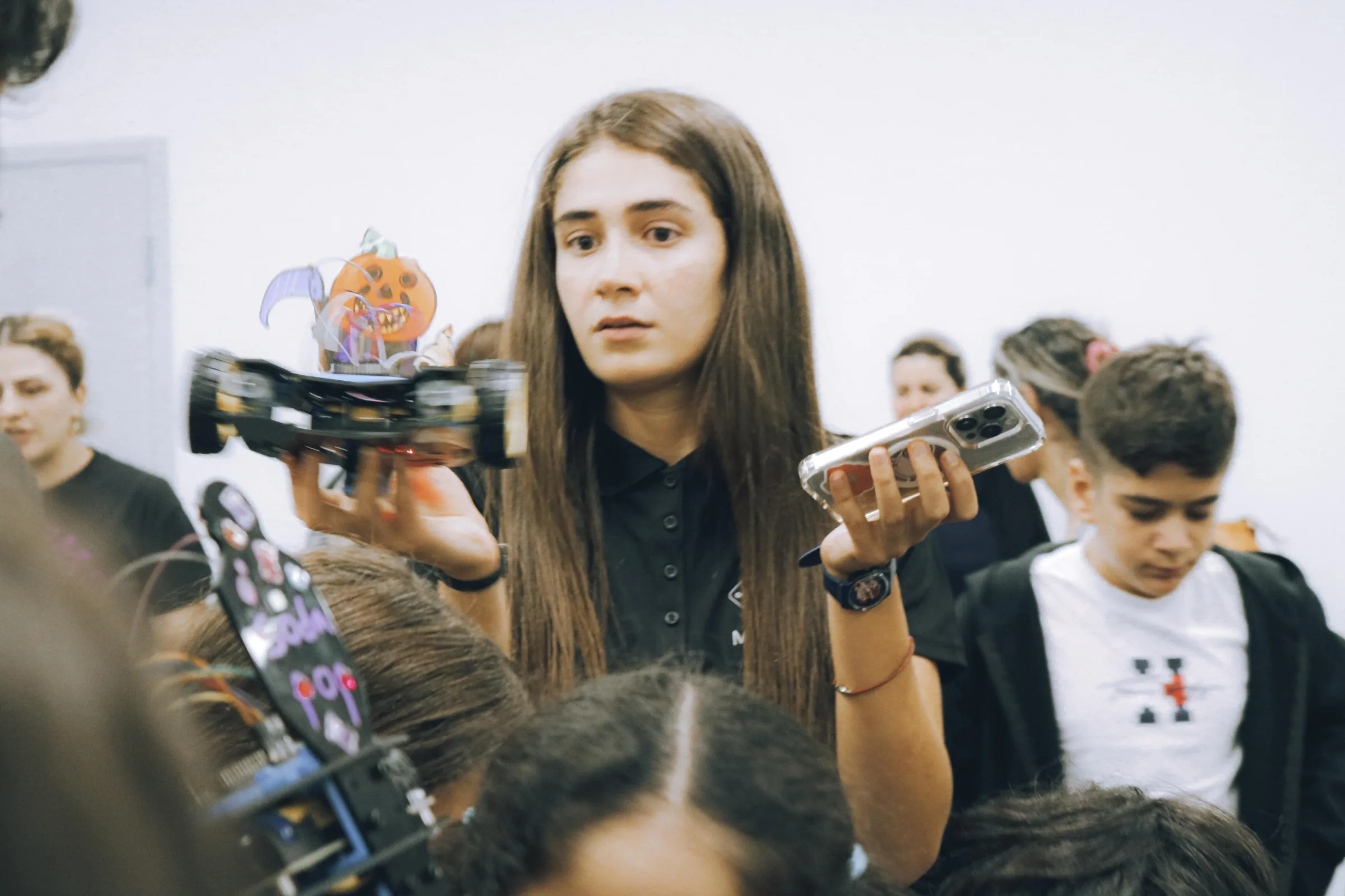 Instructor holding up a decorated robot car as students crowd around, one filming on a phone