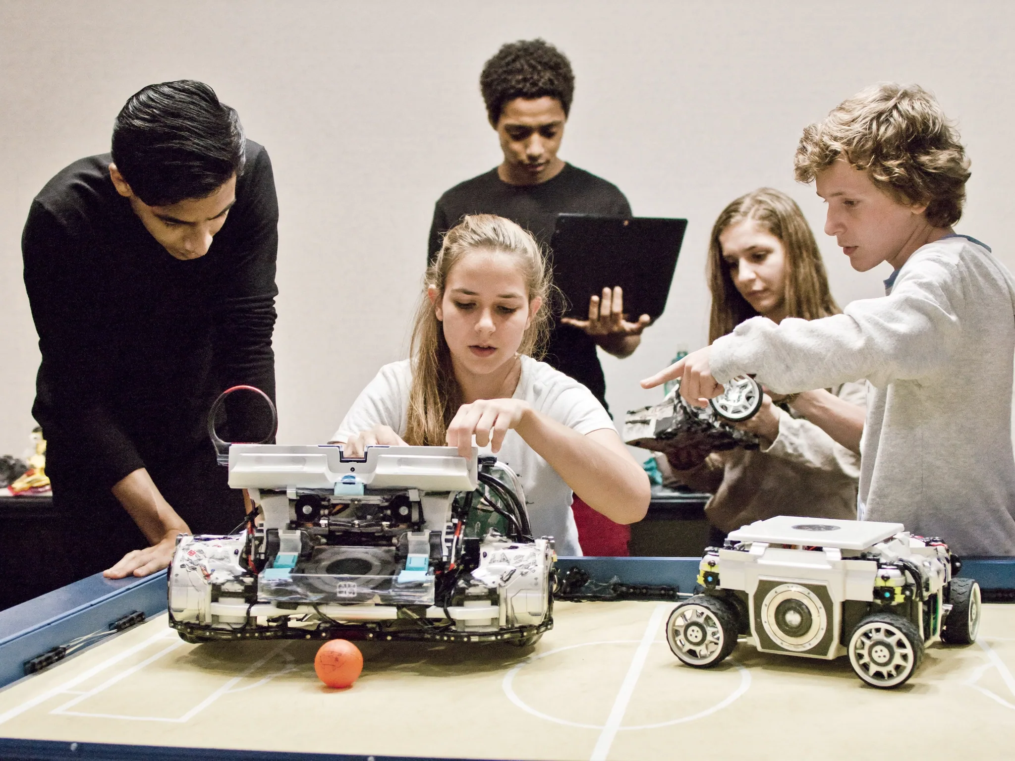 Teens adjusting two wheeled robots on a small tabletop soccer field with an orange ball