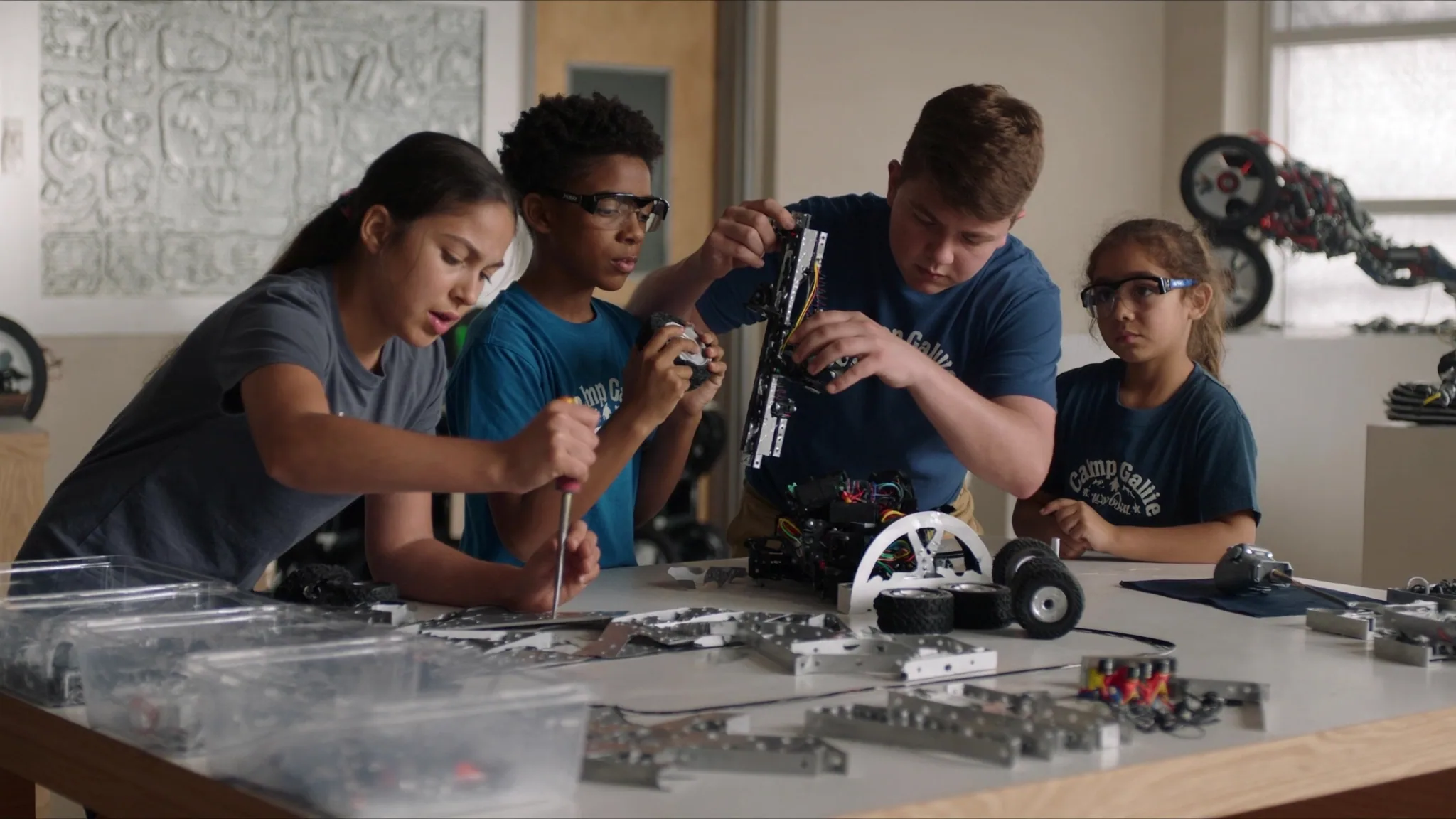 Teens in safety glasses assembling a metal robot frame with screwdrivers at a workbench