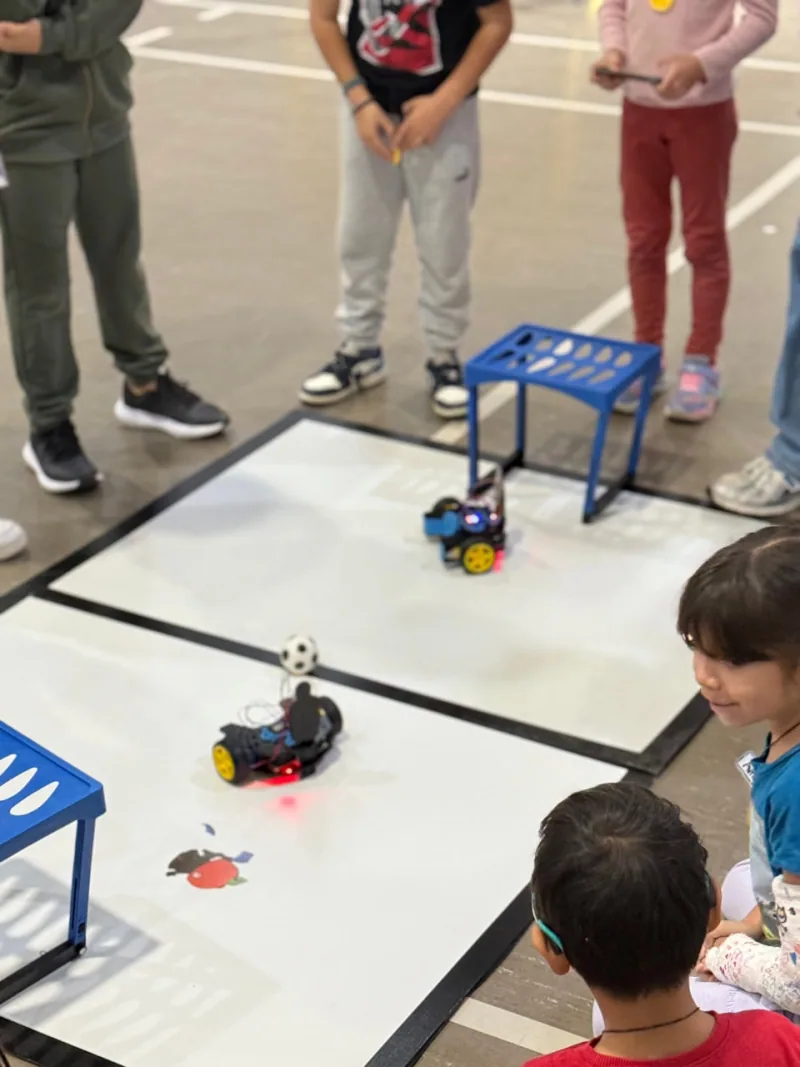 Children standing around a floor field watching two wheeled robots chase a small ball