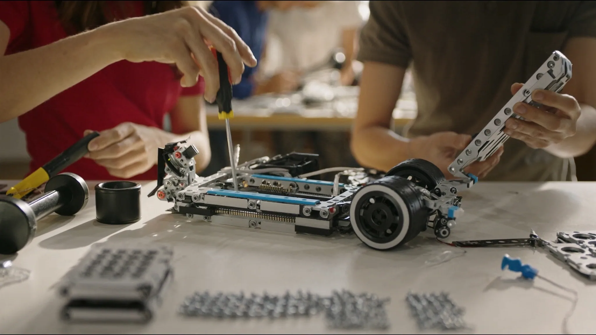 Two people fastening a metal robot chassis with screwdrivers on a parts-strewn table