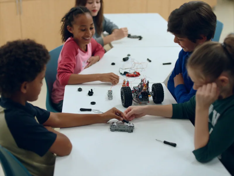 Children at a long table connecting motors and wires to a wheeled robot chassis