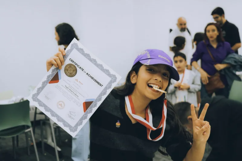 Girl wearing a medal and cap holding up a certificate of achievement and making a peace sign