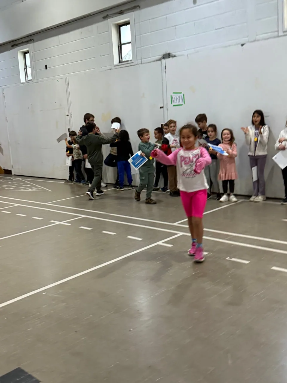 Young girl walking across a gym floor as classmates holding certificates line the wall