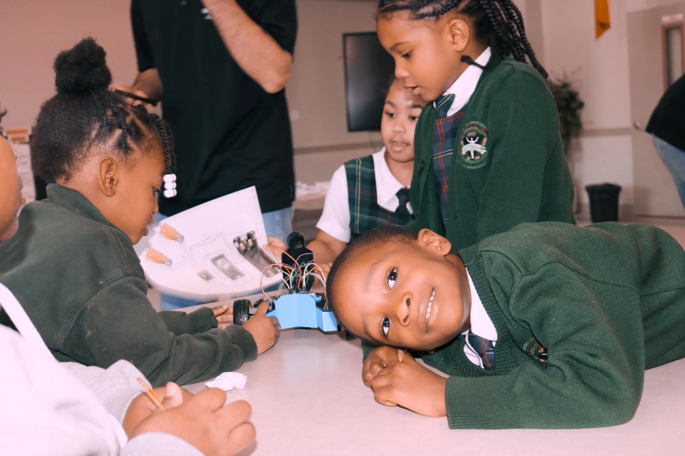 Children in school uniforms around a small robot kit, one resting his head on the table smiling