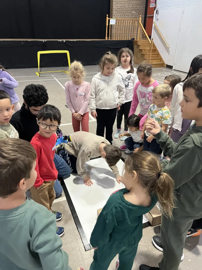 Young children crowding around a floor robot field in a gymnasium, one kneeling to look closer