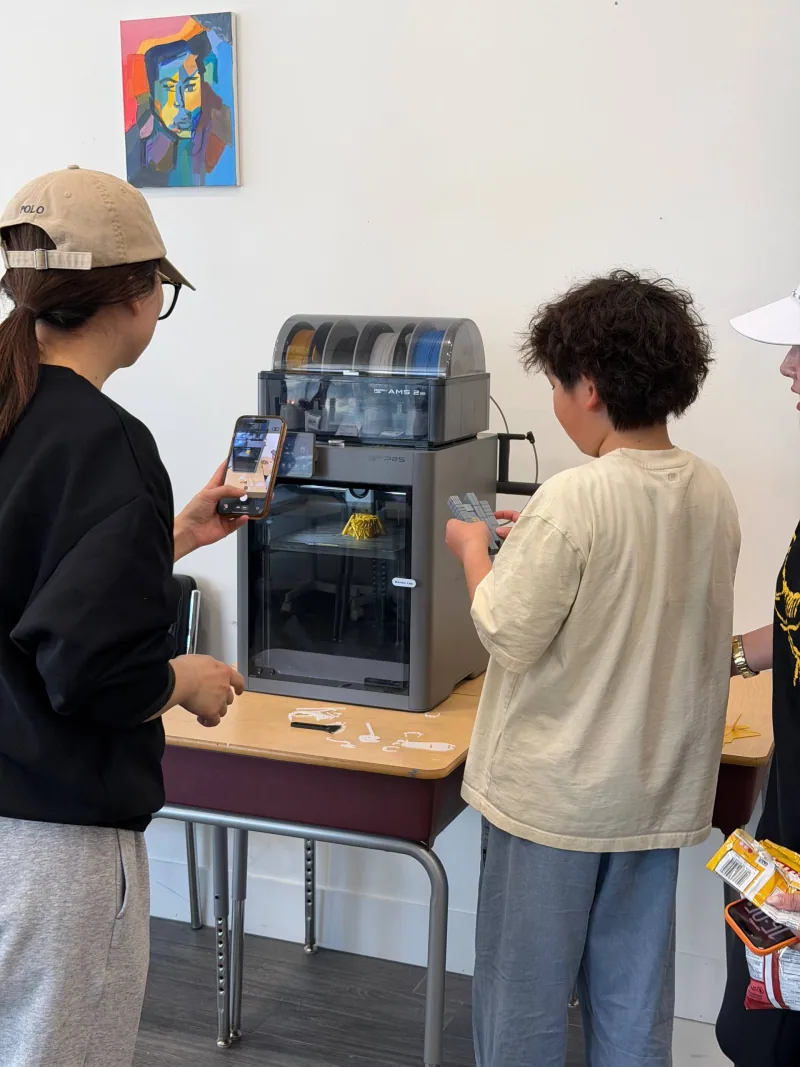 Teen photographing a 3D print in progress while another examines the printer