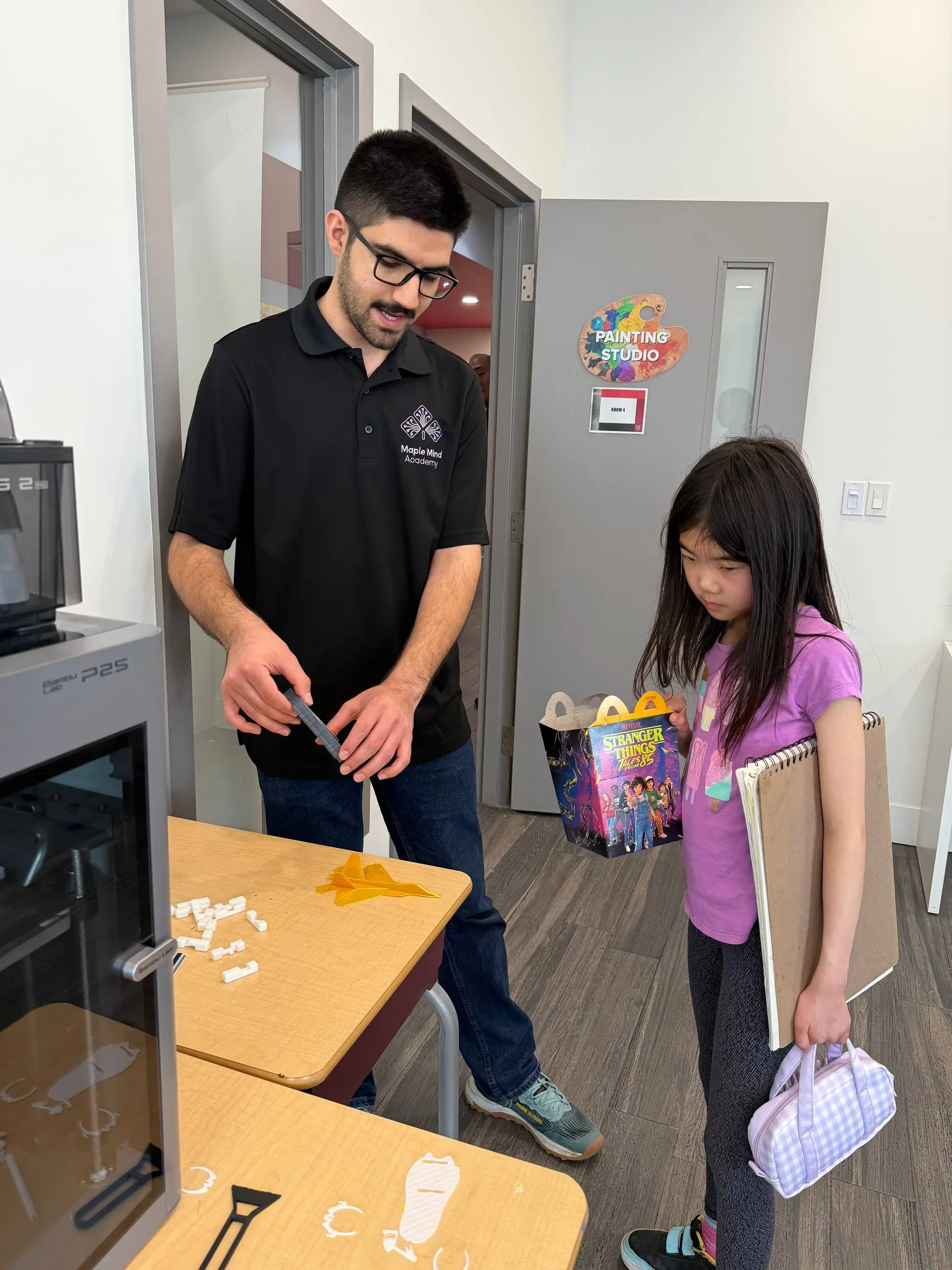 Instructor holding a 3D-printed part while a girl with a sketchbook watches beside a printer
