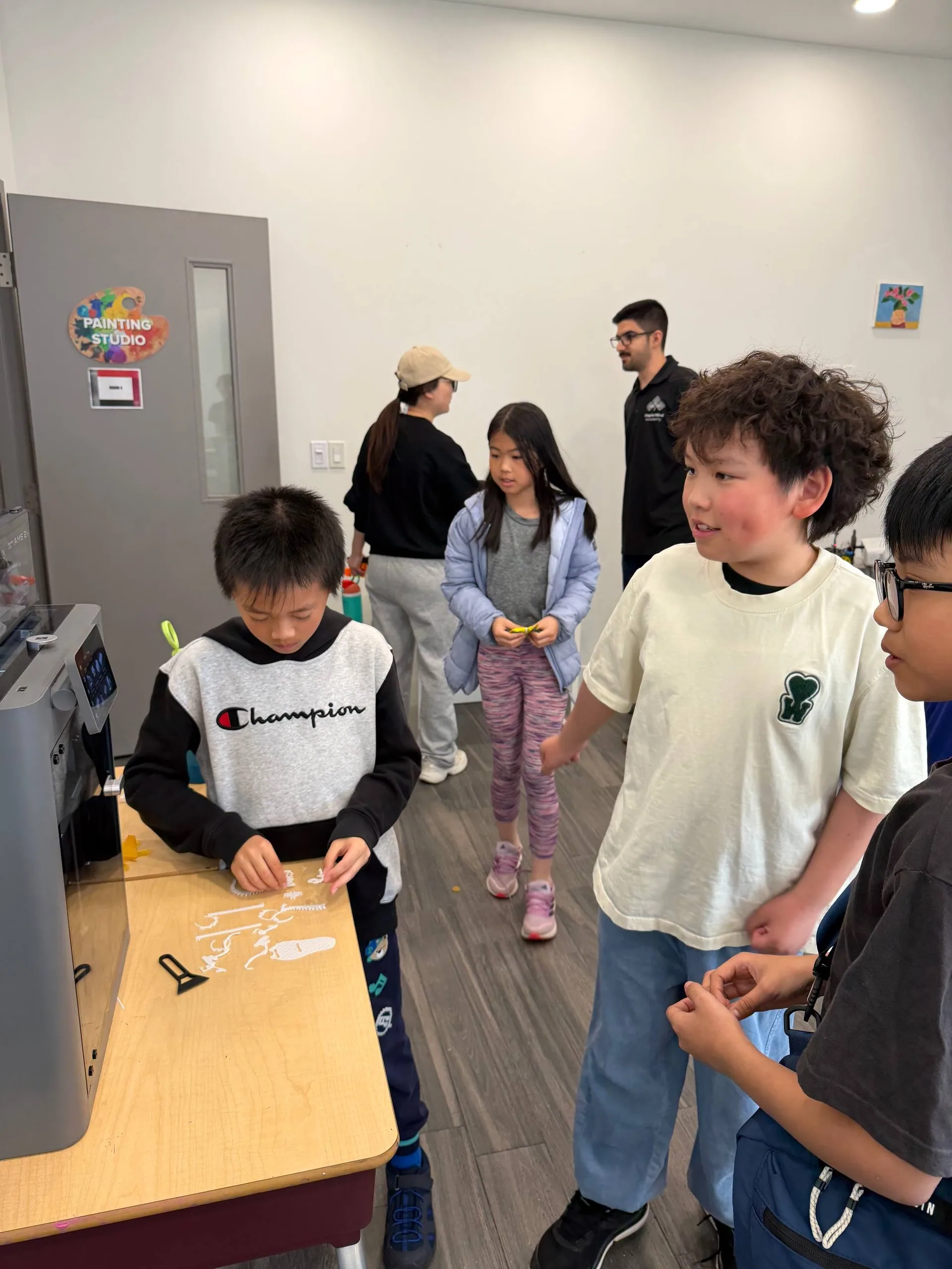 Children gathered at a desk picking up white 3D-printed pieces next to a printer