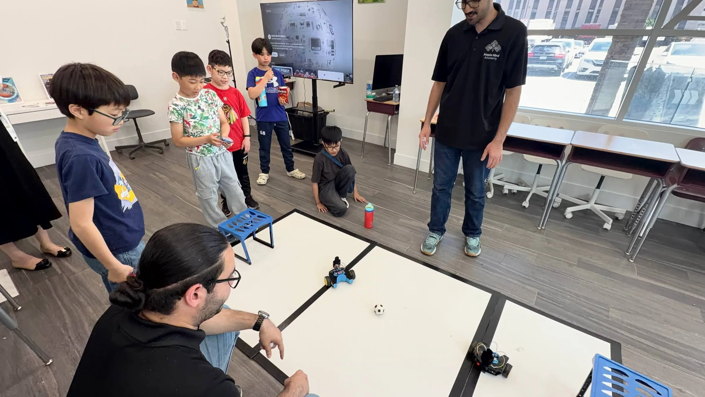 Children gathered around a floor robot soccer pitch while instructors supervise the match