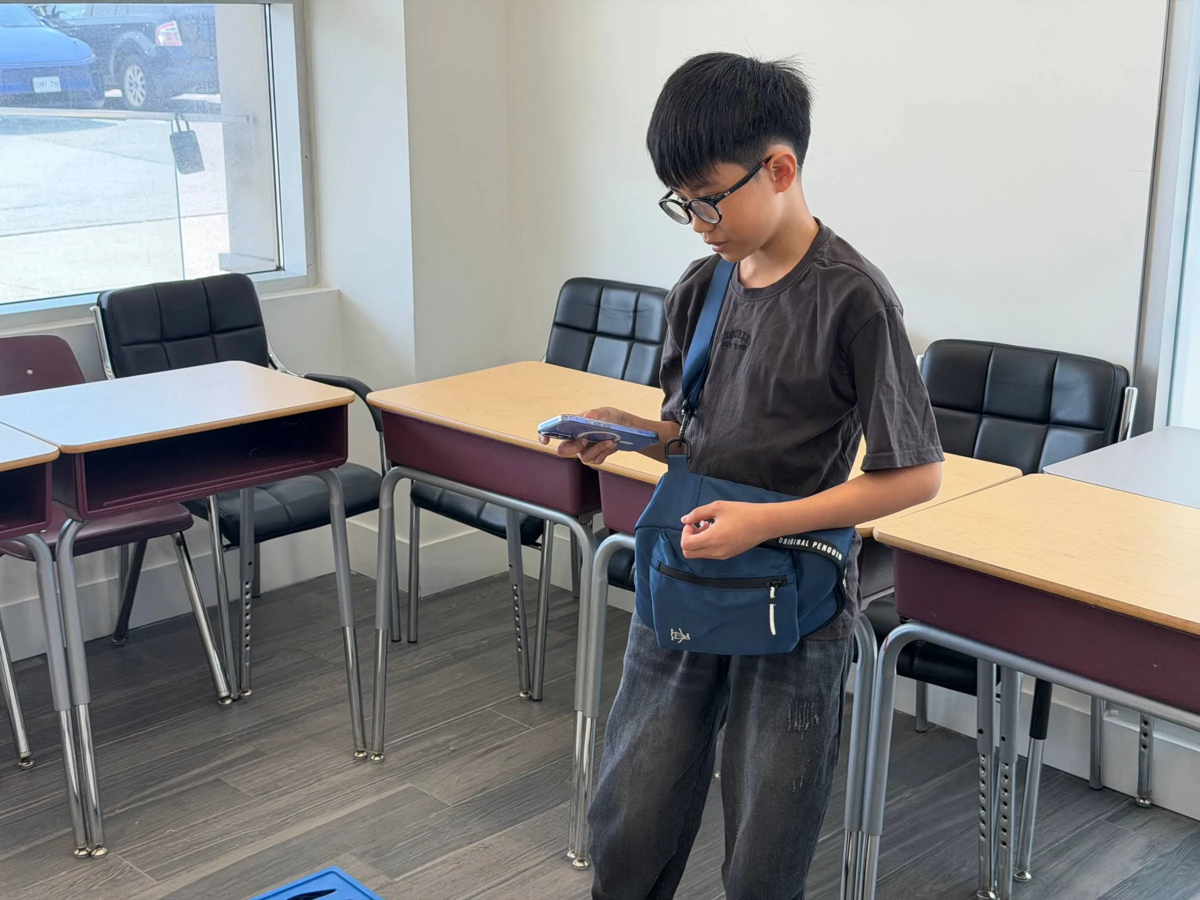 Boy in glasses focused on a phone controller while standing between classroom desks